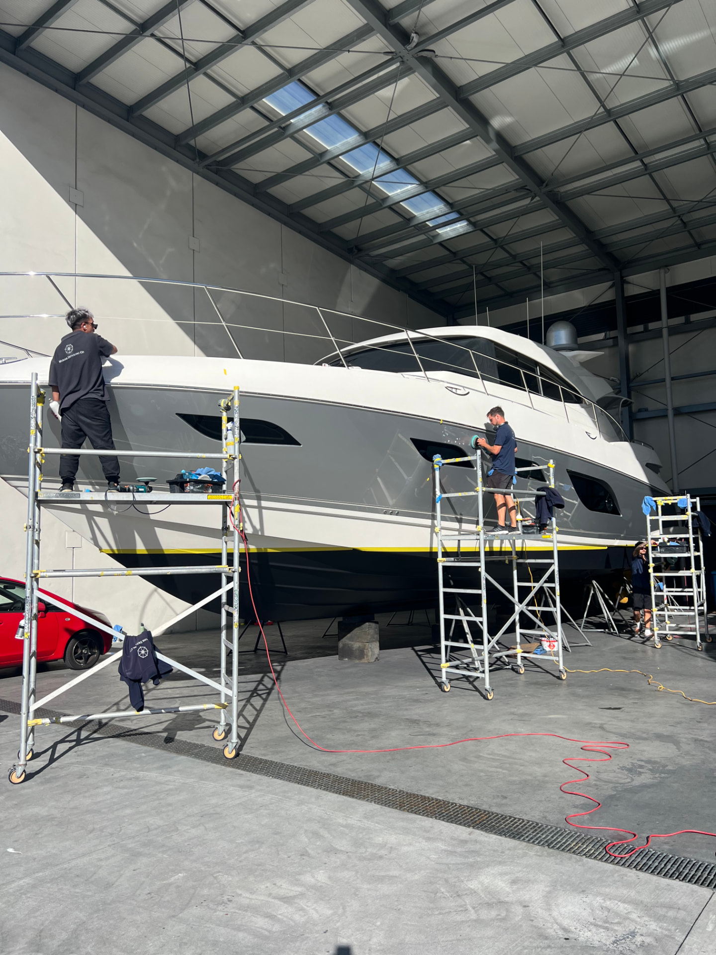 Team machine polishing a yacht hull from scaffolding inside a shed